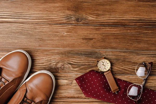 Top view of mens brown casual shoes, polka dots red tie, wristwatch and glasses on wooden background