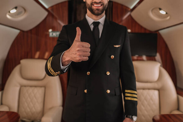 обрезанный вид улыбающегося пилота частного самолета, показывающего большой палец вверх
