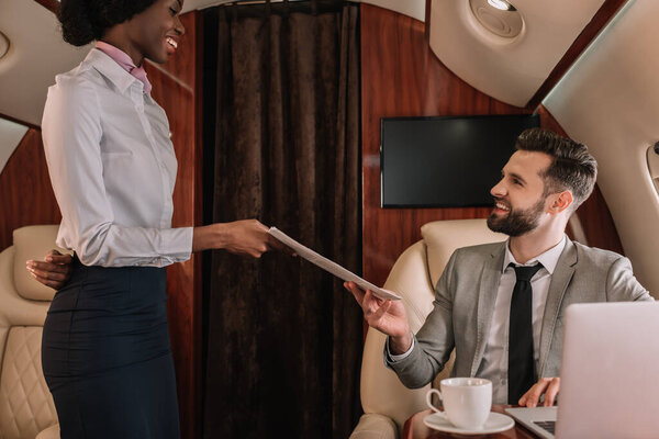 attractive african american stewardess giving newspapers to smiling businessman sitting at laptop in private jet