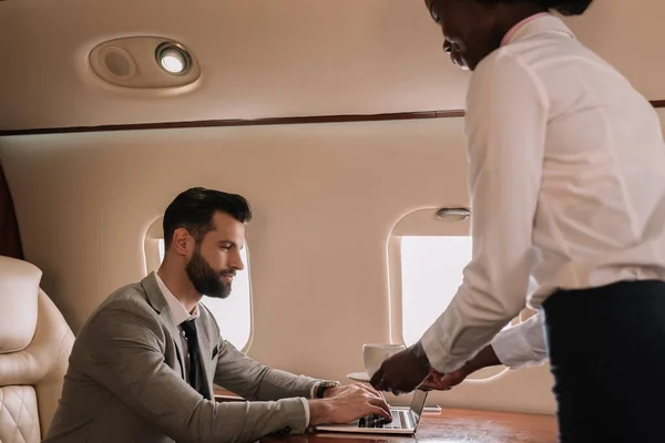 非洲裔美国空姐给在私人飞机上用笔记本电脑工作的商人一杯咖啡 — 图库照片