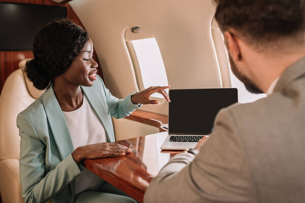 back view of businessman near smiling african american businesswoman pointing with finger and laptop with blank screen