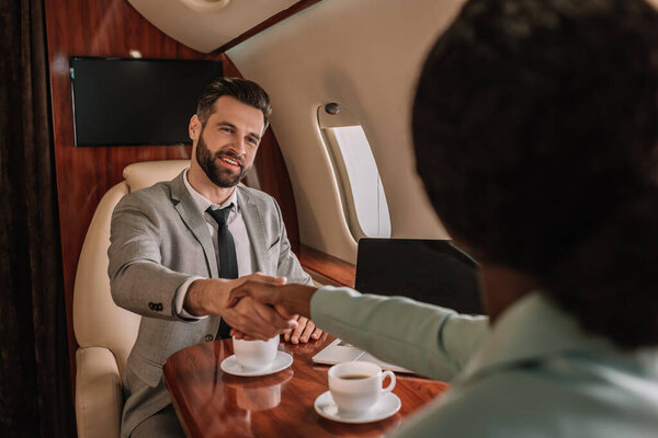 selective focus of smiling businessman shaking hands with african american businesswoman in plane