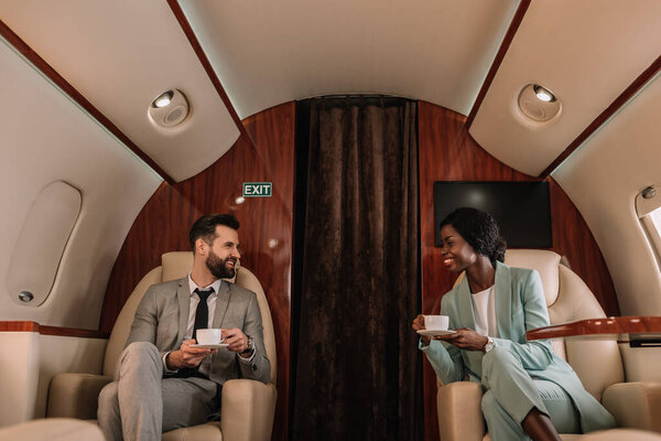 two smiling multicultural businesspeople talking while holding cups of coffee in plane