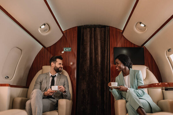 Два улыбающихся мультикультурных бизнесмена разговаривают, держа чашки кофе в самолете
