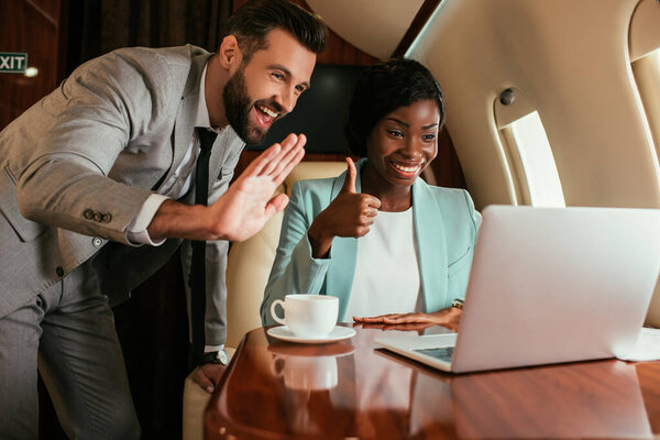 cheerful businessman waving hand and african american businesswoman showing thumb up during video call in private plane