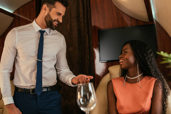 selective focus of smiling air steward looking at happy, elegant african american woman in private plane