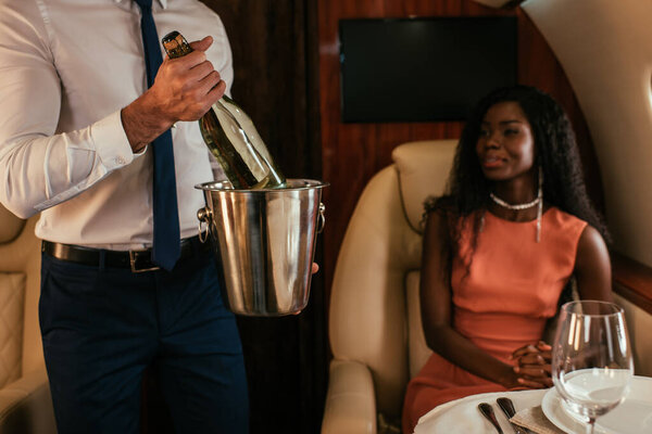 cropped view of air steward holding champagne bucket and bottle near elegant african american woman