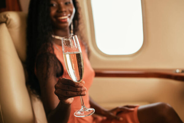 selective focus of happy african american woman looking at camera while holding glass of champagne in private jet