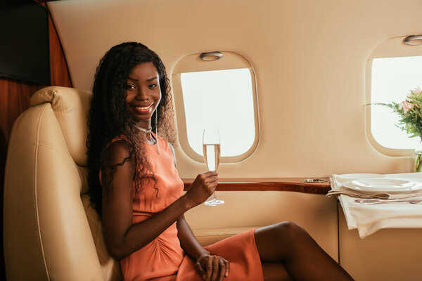 attractive african american woman hoding glass of champagne and smiling at camera while sitting near served table in private plane