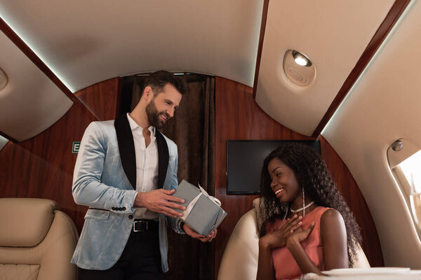 handsome, elegant man presenting gift box to suprised african american woman in private plane