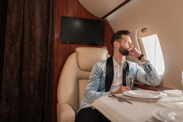 handsome, dreamy man looking in porthole while sitting at served table in private plane