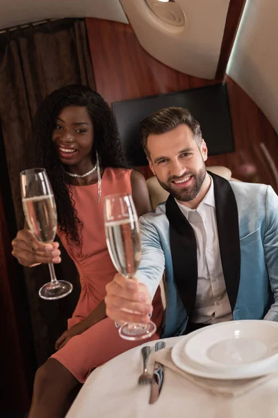 快乐优雅的跨种族夫妇一边举着香槟酒一边看着私人飞机上的相机 — 图库照片