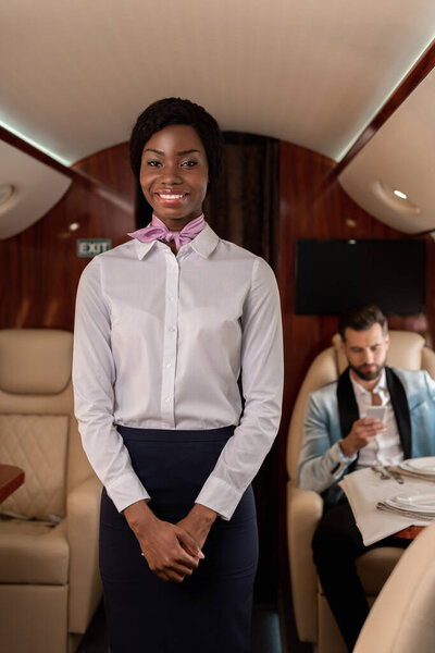 smiling african american stewardess looking at camera, while elegant man using smartphone in private plane