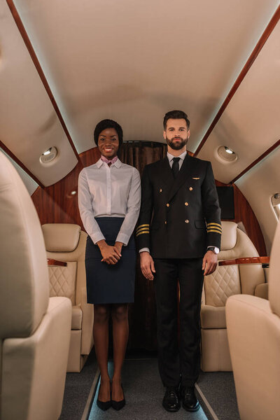 handsome, confident pilot and smiling african american stewardess looking at camera in private plane