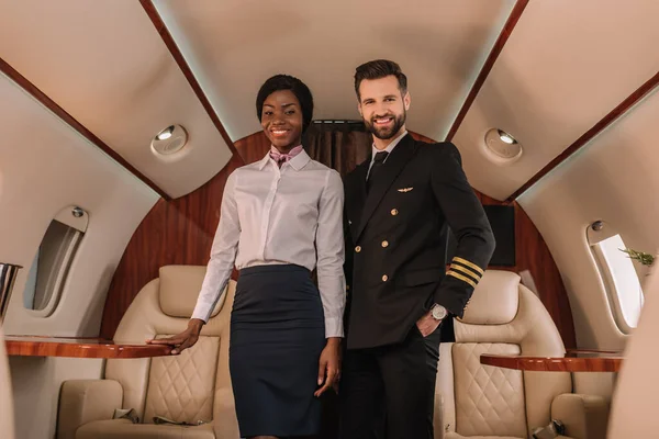 Przystojny Pozytywny Pilot Stojący Ręką Kieszeni Pobliżu Uśmiechniętej Afrykańskiej Stewardessy — Zdjęcie stockowe