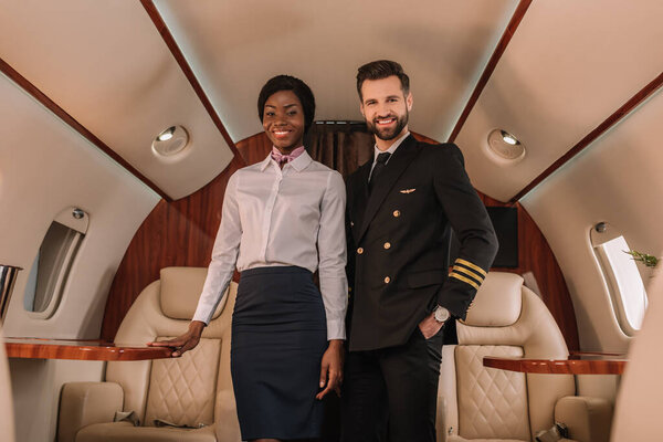 handsome, positive pilot standing with hand in pocket near smiling african american stewardess in private jet