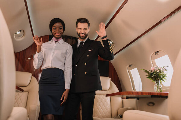 happy pilot and smiling afican american stewardess waving hands while looking at camera in private jet