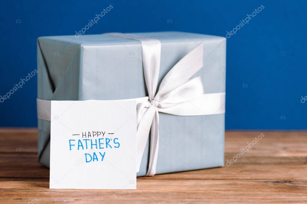 selective focus of greeting card with lettering happy fathers day and gift box with white bow isolated on blue