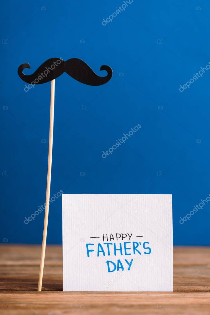 selective focus of fake mustache from cardboard and greeting card with lettering happy fathers day isolated on blue