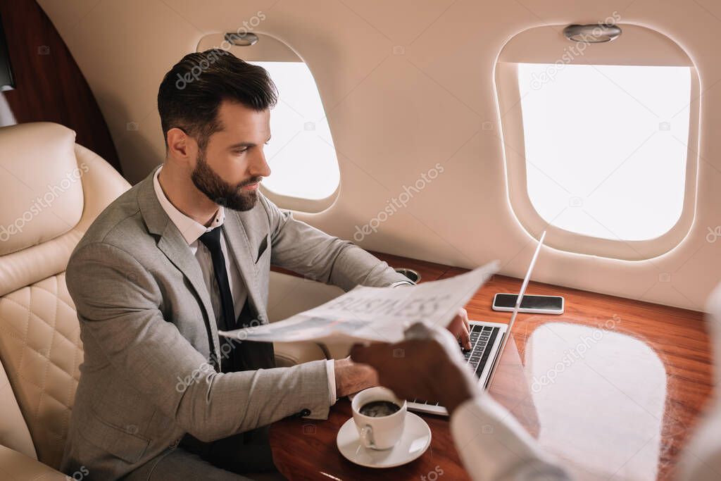 cropped view of african american stewardess giving newspaper to businessman working on laptop in private jet