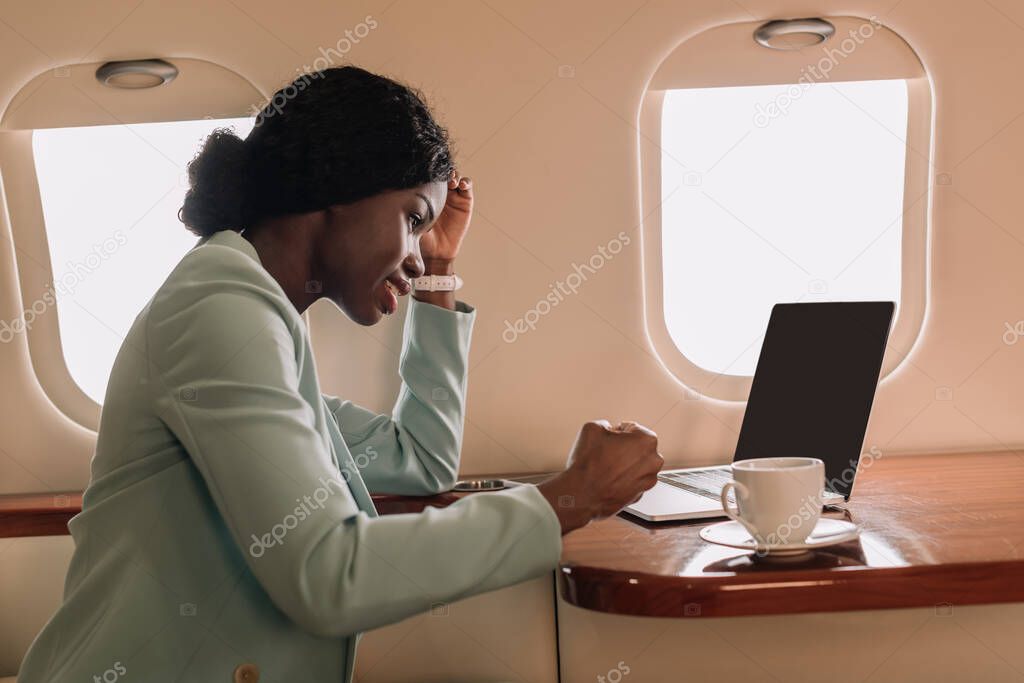 smiling african american businesswoman near laptop with blank screen and coffee cup in private plane