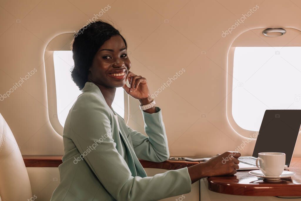 attractive african american businesswoman smiling at camera near laptop and coffee cup in private jet