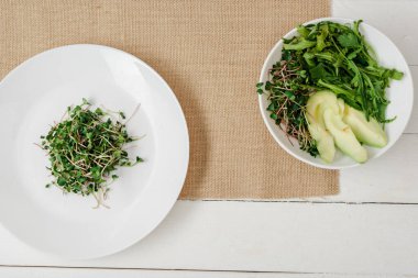 Beyaz ahşap yüzeyde bej peçetede yeşil salata kasesinin yanındaki tabakta taze mikro yeşil manzaralı.