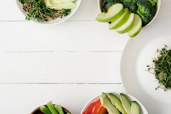 top view of fresh vegetables and fruits with microgreen in bowls on white wooden surface