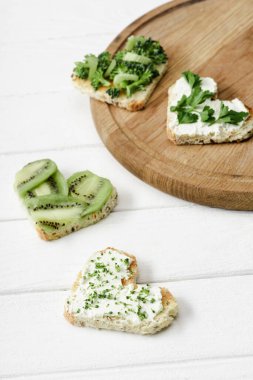 selective focus of heart shaped canape with creamy cheese, broccoli, microgreen, parsley and kiwi on board on white wooden surface clipart