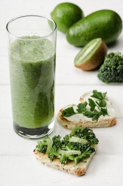 Yeşil smoothie 'nin seçici odak noktası ve krem peyniri, brokoli, meyvelerin yanında maydanoz ve beyaz ahşap yüzeyde sebze şeklinde kalp şeklinde kanepe.