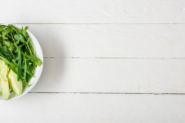 Arugula 'nın üst görüntüsü, beyaz ahşap yüzeyde avokado kasesi.