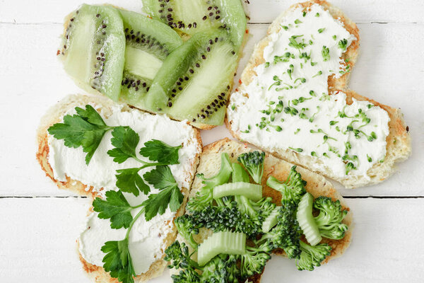 top view of heart shaped canape with creamy cheese, broccoli, microgreen, parsley and kiwi on white wooden surface