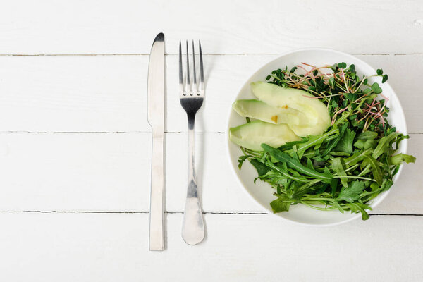 top view of arugula, avocado and microgreen in bowl on white wooden surface with fork and knife