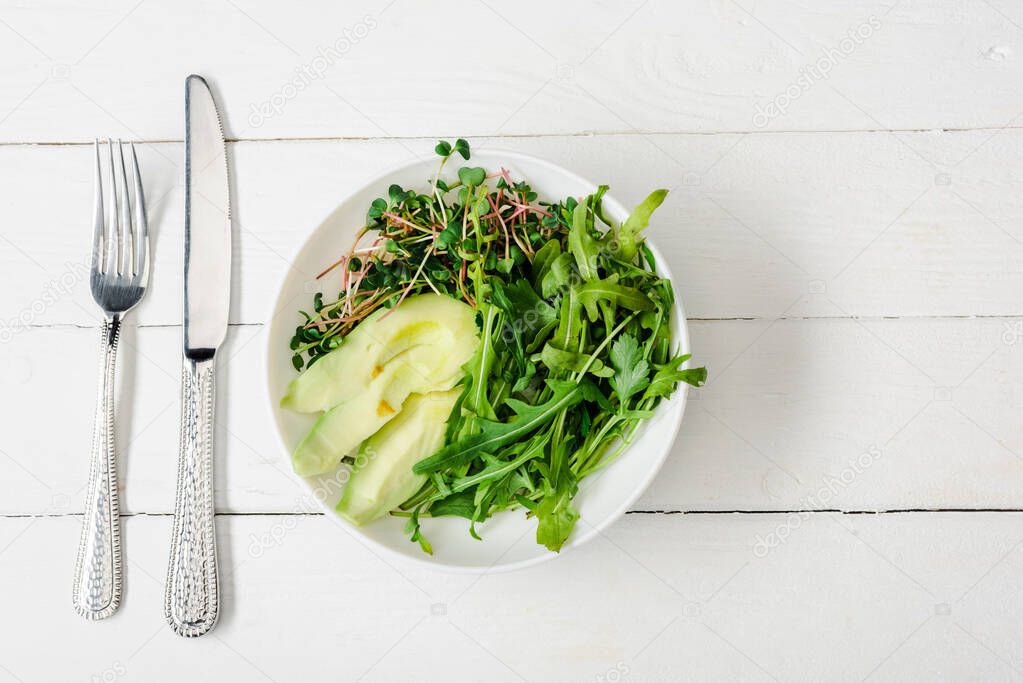 top view of arugula, avocado and microgreen in bowl on white wooden surface with cutlery