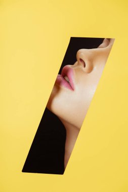 Cropped view of female face with pink lips in quadrilateral hole in yellow paper on black clipart