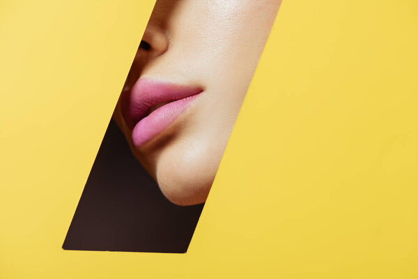 Partial view of female face with pink lips in quadrilateral hole in yellow paper on black
