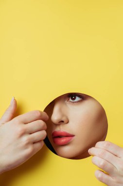 Woman with coral lips looking across round hole and touching yellow paper with hands  clipart