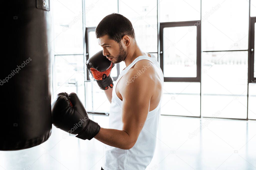 side view of handsome sportsman in boxing gloves training with punching bag in sports center 