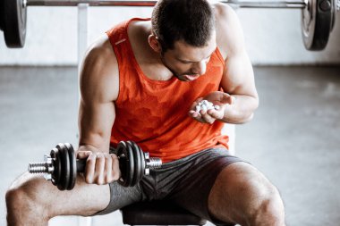 Ağzı açık sporcu, halterle egzersiz yapıyor ve spor salonunda probiyotik haplara bakıyor.