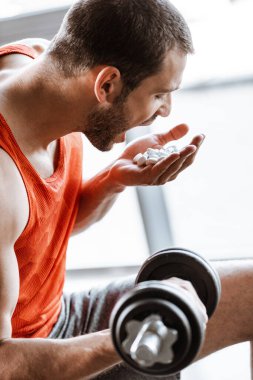 Ağzı açık, dambılla egzersiz yapan ve spor salonunda probiyotik haplara bakan sporcunun seçici odağı.