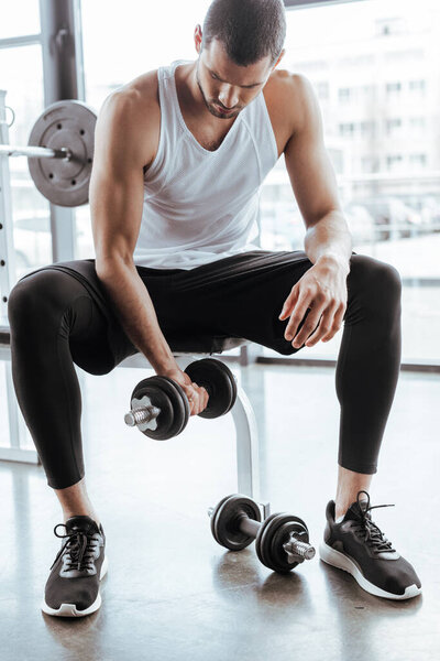 handsome sportsman exercising with dumbbell while sitting in gym 
