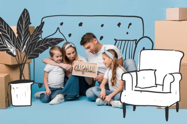 positive parents and kids holding card with home sweet home lettering and furniture illustration while sitting on blue with cardboard boxes for relocation clipart
