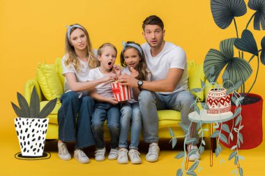 şaşırmış bir aile kanepede film izliyor. Sarı üzerinde patlamış mısır kovası, bitkiler resimli.