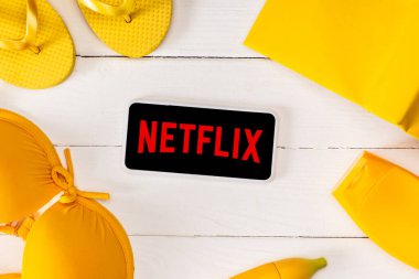 KYIV, UKRAINE - 25 Mart 2020: Beyaz ahşap yüzey üzerinde netflix uygulaması olan güneş kremi, mayo ve kitap ile sarı parmak arası terliklerin en üst görüntüsü