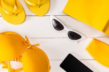 Güneş gözlüklerinin üst görüntüsü, güneş şapkalı sarı mayo ve beyaz ahşap yüzeyde akıllı telefon.