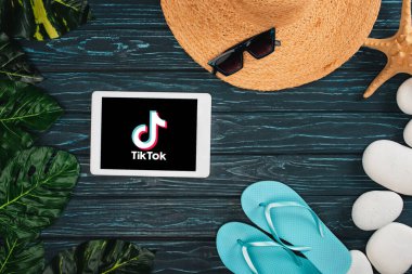 KYIV, UKRAINE - MARCH 25, 2020: Top view of digital tablet with TikTok app near flips flops, straw hat with pebbles and leaves on dark wooden surface clipart