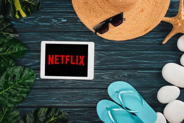 KYIV, UKRAINE - 25 Mart 2020: Hasır şapka, parmak arası terlik ve koyu ahşap yüzey üzerinde yapraklar ile netflix uygulaması ile dijital tabletin üst görünümü