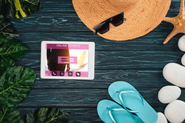 Dijital tabletin en üst görüntüsü online bilet uygulaması hasır şapka, parmak arası terlik ve koyu ahşap yüzey üzerindeki deniz taşları