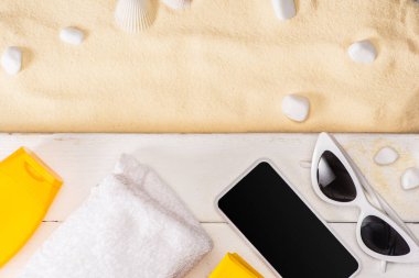 Akıllı telefonun üst görüntüsü, havluyla güneş gözlüğü ve beyaz tahta üzerinde güneş kremi ve kumdaki çakıl taşları.