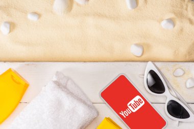 KYIV, UKRAINE - MARCH 25, 2020: Top view of smartphone with youtube app near sunglasses, sunscreen and towel on white wooden planks and pebbles on sand  clipart